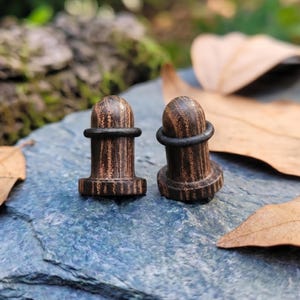 May include: Two wooden ear plugs with a dark brown finish and a black rubber ring. The plugs have a cylindrical shape with a rounded top and a flared base. The wood grain is visible, adding texture to the plugs. They are displayed on a blue-grey stone surface.