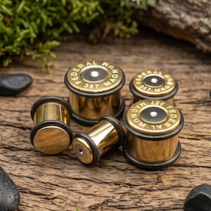 May include: A collection of five gold-colored ear plugs designed to resemble 357 Magnum bullet casings. Each piece features a metallic brass-toned body with a black rubber O-ring and a flat top stamped with the text FEDERAL MAGNUM 357.