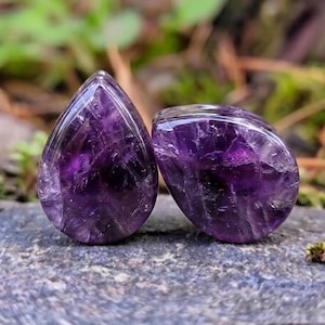 May include: Two teardrop-shaped ear plugs crafted from polished deep purple amethyst stone. Each piece features a smooth, rounded surface with visible natural crystalline inclusions and variations in the dark violet colour throughout the stone.