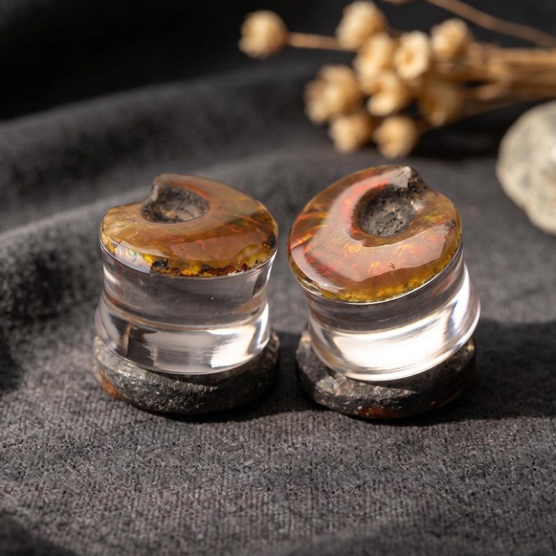 May include: A pair of ear plugs featuring a clear, transparent cylindrical body with a dark, textured stone base and a polished, iridescent amber-colored top surface that displays a crescent-shaped indentation on the upper edge of each piece.