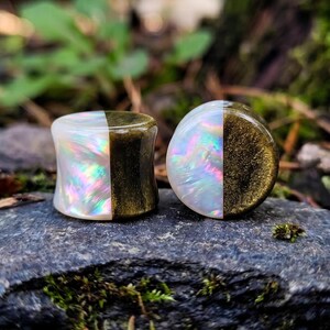 May include: A pair of circular ear plugs featuring a split design. One half of each plug is made of a white, iridescent material with a pearlescent sheen, while the other half is a dark, metallic gold with a fine, shimmering glitter texture.