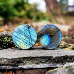 Puede incluir: Dos tapones redondos de piedra labradorita. Los tapones muestran un vibrante juego de colores, con azules, verdes y toques de amarillo. Las piedras tienen una superficie pulida y están sobre un fondo natural.