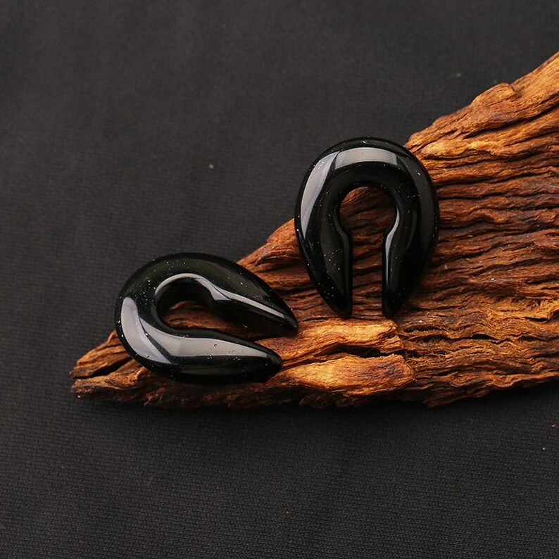 May include: Two black horseshoe-shaped ear plugs with a smooth, glossy finish and subtle glittery flecks. These jewelry pieces feature a curved, open design, showcasing a polished surface that reflects light, presented as a pair of body jewelry.