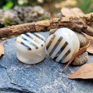 Puede incluir: Un par de expansores de oreja circulares con una base blanca iridiscente lisa y rayas diagonales incrustadas de labradorita oscura y brillante. Los expansores tienen un acabado pulido que refleja la luz, resaltando el contraste entre el material nacarado y las bandas minerales oscuras de tonos tornasolados.