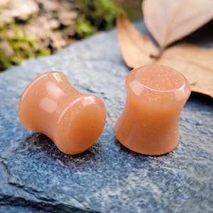 Puede incluir: Dos tapones para los oídos de color naranja con una ligera forma de reloj de arena. Los tapones tienen una superficie lisa y pulida con sutiles inclusiones brillantes. Se colocan sobre una superficie de piedra azul-gris texturizada, con un fondo natural borroso.