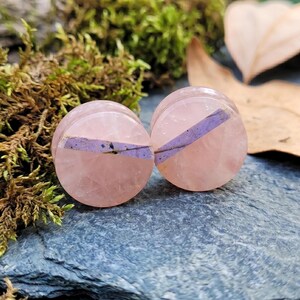 Puede incluir: Un par de expansores de oreja circulares hechos de piedra rosa pálido translúcida, cada uno con una incrustación diagonal distintiva de material morado claro que atraviesa el centro de la superficie lisa y pulida de las piezas de joyería.