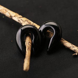 May include: A pair of black, glossy, teardrop-shaped ear gauges with a smooth, polished finish. Each piece features a central opening and a tapered point at the bottom, designed for body modification jewelry, resting on a textured wooden twig.