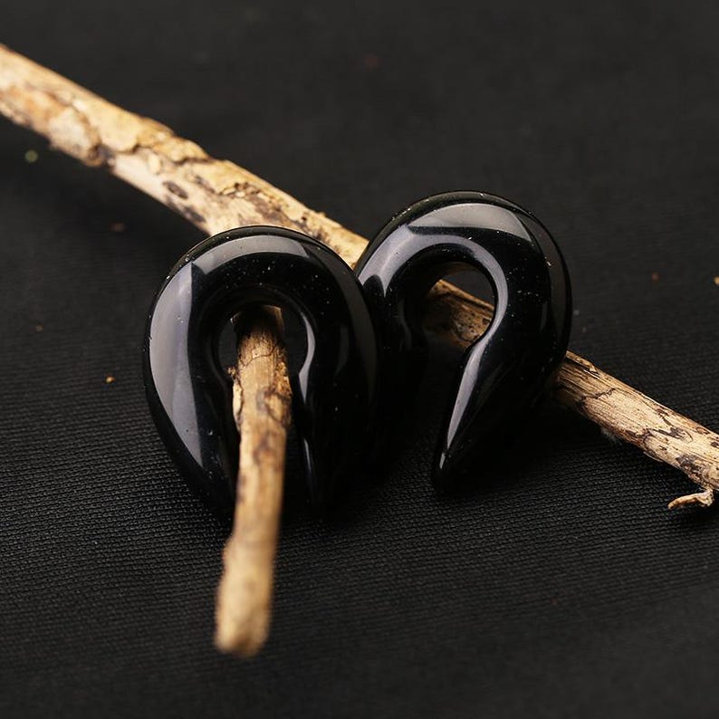 May include: A pair of black, glossy, teardrop-shaped ear gauges with a smooth, polished finish. Each piece features a central opening and a tapered point at the bottom, designed for body modification jewelry, resting on a textured wooden twig.