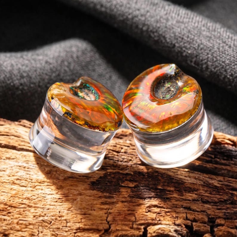 May include: A pair of circular ear plugs featuring clear, transparent bases and tops inlaid with iridescent, amber-toned material displaying flashes of green, orange, and red light, creating a vibrant, multi-colored effect within the resin.