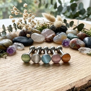 May include: Five barbell-style body jewelry pieces with round, cat's eye stone cabochon ends in light green, white, light blue, pink, and orange. Each piece features a metallic post and a spherical backing, arranged in a row on a wooden surface.