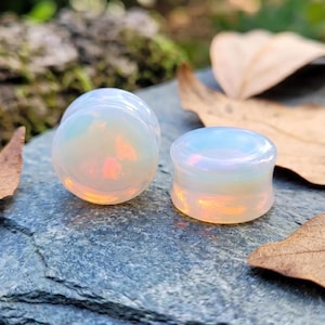 Puede incluir: Dos tapones de ópalo iridiscente, que muestran un degradado de color blanco nacarado y naranja. Estos expansores de oreja están diseñados con una forma cilíndrica redondeada. Los tapones se colocan sobre un fondo natural de piedra y follaje.