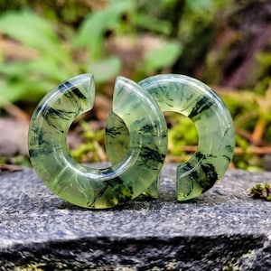 May include: A pair of C-shaped ear gauges crafted from translucent pale green stone featuring dark, moss-like dendritic inclusions throughout the material. The circular jewelry pieces have a smooth, polished finish and a gap at the top.