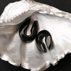 May include: Two black, U-shaped ear gauges with a smooth, glossy finish. These teardrop-shaped body jewelry pieces feature an open gap at the top and are displayed resting inside a large, silver-toned, textured seashell with a metallic sheen.