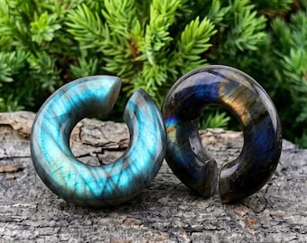 May include: A pair of circular ear weights crafted from polished labradorite stone. The left piece displays a vibrant blue flash, while the right piece features deep blue and gold tones. Both items have a small opening along the ring.