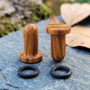 Puede incluir: Dos dilatadores de oreja cilíndricos de madera con acabado pulido y vetas de madera visibles. Cada pieza presenta una cara circular plana y un extremo redondeado, acompañados por dos juntas tóricas de goma negra independientes.