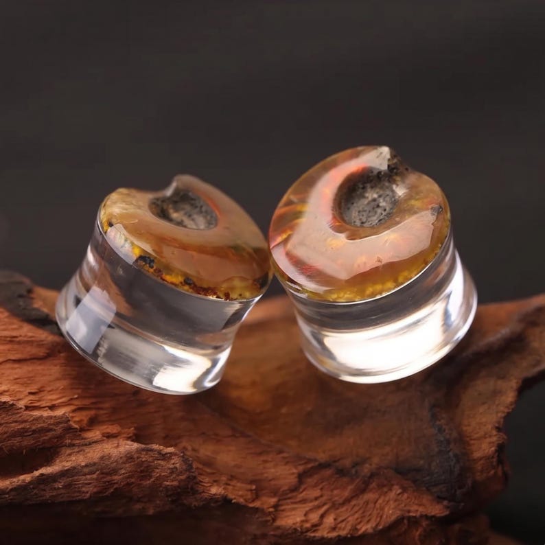 May include: A pair of circular ear plugs featuring clear, transparent sides and a top surface with a polished, amber-colored resin inlay. The inlay displays a swirling, organic pattern with deep orange and brown tones surrounding a dark, textured center.