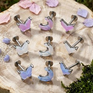 Puede incluir: Una colección de nueve piercings labret de metal con adornos en forma de mariposas, medias lunas y corazones. Los ornamentos están fabricados con un material translúcido y opalescente en tonos rosa, blanco y azul.