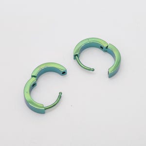 Puede incluir: Un par de pendientes de aro pequeños con un acabado metálico iridiscente que cambia entre tonos verdes y azules. Cada pendiente cuenta con un mecanismo de cierre de bisagra con un poste fino que encaja en un pequeño orificio lateral.
