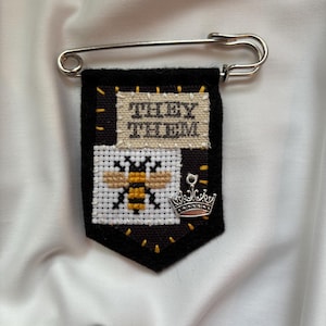Op de afbeelding: Een zwarte stoffen badge bevestigd aan een zilveren veiligheidsspeld. De badge bevat een kruissteek patch met de tekst "THEY THEM" en een tweede patch met een bij. Een kleine zilveren kroonbedel is aan de rechteronderkant bevestigd.