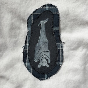 Puede incluir: Parche de tela con un diseño de murciélago gris sobre un óvalo negro. El murciélago está colgado boca abajo. El óvalo negro está rodeado de tela a cuadros azul y negro, con un borde deshilachado. Sobre un fondo blanco.