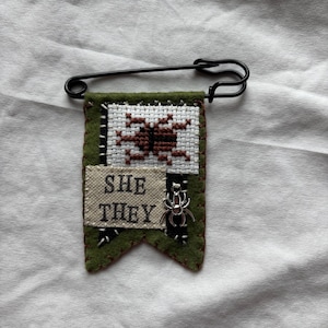 May include: A green felt pennant attached to a large black safety pin. The pennant features a white cross-stitched square with a brown beetle, a beige fabric patch with the black text "SHE THEY", and a small silver-toned metal spider charm.