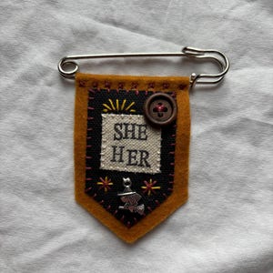 May include: A mustard yellow felt pennant pin featuring a black fabric center with the text "SHE HER" embroidered in white. The design includes a brown button, yellow sunburst stitching, pink star accents, and a silver broomstick charm.