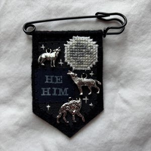 May include: A black felt pennant attached to a large black safety pin. The pennant features an embroidered gray moon, three silver-toned wolf charms, and a dark blue patch with the embroidered white text HE HIM, accented by small white cross stitches.