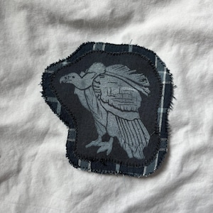 May include: A dark gray patch featuring a detailed illustration of a vulture. The patch is sewn onto a dark blue fabric with a plaid pattern around the edges. The background is a white fabric.