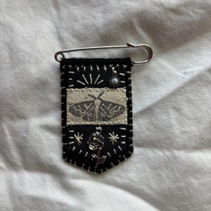 May include: A black fabric pin with a moth design, white stitching, and a silver safety pin. The pin features a small, silver rose and a decorative bead. The moth design is in a rectangular patch.