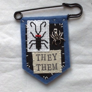 May include: A blue felt pennant attached to a black safety pin. The pennant features a white cross-stitched insect with a red collar, a silver spider charm, and a beige fabric patch with the black embroidered text THEY THEM.