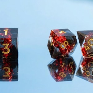 Può includere: Tre dadi poliedrici realizzati in resina scura traslucida con scaglie metalliche rosse incorporate e numeri incisi color oro. I dadi sono disposti in fila e si riflettono su una superficie liscia, includendo un cubo, un d8 e un d10.