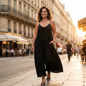 Op de afbeelding: Een persoon in een zwarte jumpsuit met spaghettibandjes en zakken loopt door een stadsstraat. Ze draagt zwarte sandalen en gouden armbanden. De achtergrond toont een Europese stad met cafés en winkels.