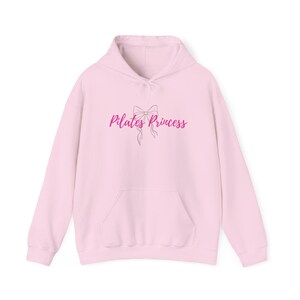 Peut inclure: Un sweat-shirt à capuche rose clair avec une poche avant et un cordon de serrage. Le texte "Pilates Princess" est imprimé en cursive avec un motif de nœud. Le sweat-shirt est fait d'une matière douce.