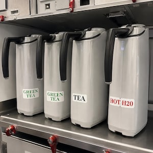 May include: Four gray insulated beverage dispensers with black handles are lined up on a stainless steel shelf. Labels read "GREEN TEA", "TEA", and "HOT H2O". The dispensers are in a commercial setting.