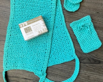 May include: A set of crocheted bath accessories in a turquoise colour. The set includes a long bath scrub, two small round scrubbers, and a small rectangular soap dish. The soap dish has a bar of soap inside with the text "Coconut" and "Refreshing Vegetable Soap" on the label.