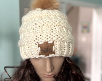 May include: Cream-colored knit beanie with a cable knit design and a faux fur pom-pom. The hat features a brown leather patch with the text "sahs". This winter hat is a cozy accessory.