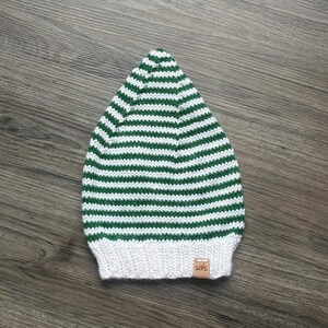 May include: A knitted beanie featuring a pattern of horizontal green and white stripes. The hat has a solid white ribbed brim at the base and a small, rectangular tan leather-like tag with the text "sahs" stitched onto the lower right side.