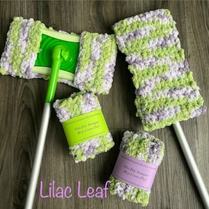 May include: A set of handmade, reusable, machine washable wet/dry sweeper mop cover pads in a lilac leaf color scheme. The mop covers are a mix of green and lavender yarn. The image also shows a green and silver mop handle.