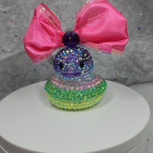May include: A rubber duck figurine covered in multi-colored iridescent rhinestones, featuring long black eyelashes and a large, bright pink satin bow attached to the top of its head with a dark purple circular gem at the center of the bow.