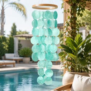 May include: A hanging wind chime featuring multiple translucent, circular turquoise discs suspended from a light-coloured wooden ring. The discs are arranged in vertical strands, creating a layered, cascading effect that catches the light.