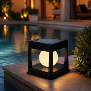 Op de afbeelding: Een vierkante zwarte buitenlantaarn met een centrale gloeiende bolvormige lamp. Het armatuur heeft een open frameontwerp met een plat bovenoppervlak en een stevige basis, die een warm en zacht licht uitstraalt naar de omgeving.
