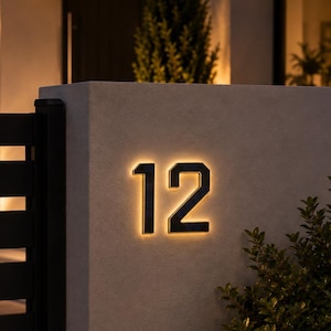 Könnte beinhalten: Ein Hausnummernschild mit den Ziffern 12 in einer dunklen, kantigen Schriftart. Die Zahlen sind an einer hellen Wand montiert und verfügen über eine warme, leuchtende Hintergrundbeleuchtung, die einen sanften Lichtschein erzeugt.