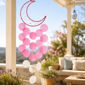 May include: A hanging mobile featuring a crescent moon frame wrapped in pink thread with a webbed centre. Suspended from the moon are multiple translucent pink circular discs arranged in a cluster, followed by three clear circular discs at the bottom.