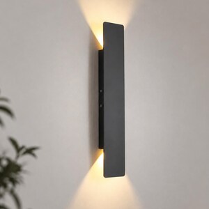 Op de afbeelding: Een verticale rechthoekige wandlamp met een matzwarte afwerking. Het armatuur heeft een langwerpig, plat ontwerp dat warm, gloeiend licht uitstraalt vanaf zowel de boven- als onderrand, waardoor twee duidelijke lichtbundels op de muur ontstaan.