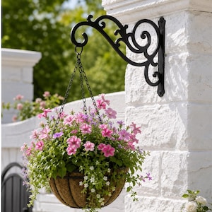 Könnte beinhalten: Eine schwarze Wandhalterung aus Metall mit Schnörkeln an einem weißen Ziegelpfeiler, die einen Blumenampel mit rosa und weißen Blüten trägt. Der Korb ist an drei dünnen schwarzen Ketten befestigt, die an dem geschwungenen Metallarm hängen.