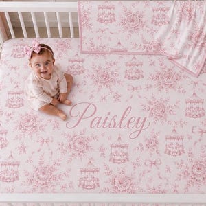 Op de afbeelding: Een wieglaken en bijpassende deken met een roze en wit toile-patroon met carrousel- en bloemmotieven. Het laken is gepersonaliseerd met de naam Paisley in cursief schrift. De set wordt getoond in een wit houten ledikant.