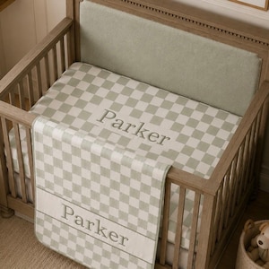 May include: A wooden crib featuring a sage green and white checkered blanket and matching crib sheet. Both the blanket and the sheet are personalized with the name Parker embroidered in a light gray font across a solid horizontal band.