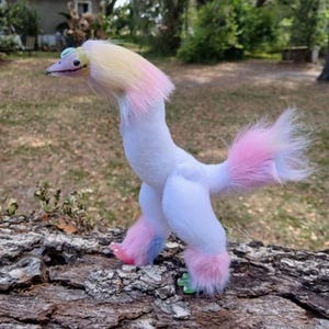 May include: A whimsical, handmade plush bird with a long neck and fluffy tail. The bird is primarily white, with pink, yellow, and blue accents on its head, tail, and feet. It stands on a piece of wood, showcasing its unique design.
