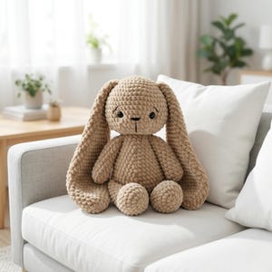 以下が含まれることがあります： 長い垂れ耳と優しい表情の、ベージュ色のウサギのぬいぐるみ。手作りのぬいぐるみは白いソファに座っています。ウサギは織り目加工の外観で、柔らかいおもちゃです。