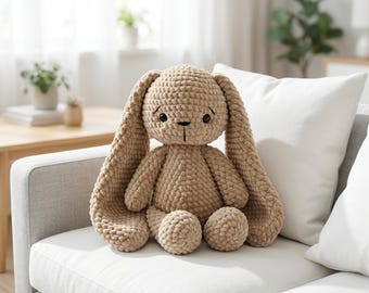 May include: A plush, tan-colored bunny rabbit with long, floppy ears and a sweet expression. The handmade stuffed animal is sitting on a white couch. The bunny has a textured, knitted appearance and is a soft toy.